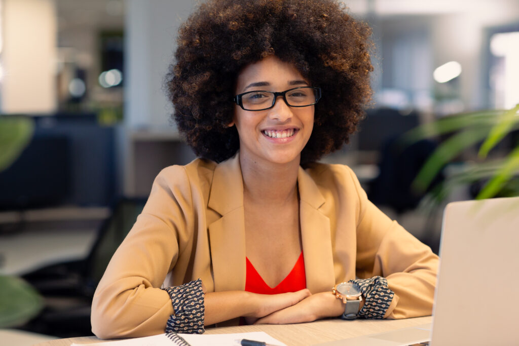 Portrait of smiling african american businesswoman sitting with laptop at workplace. unaltered, business, modern office and technology concept.